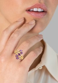 Goldring mit drei lila Edelsteinen, mit einem gewellten Design. Hand in der Nähe des Gesichts vor einem hellen Hintergrund platziert.