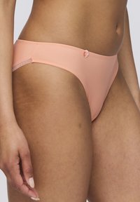 Peach-colored thong with smooth texture, low-rise design, and a small decorative button on the side. Elasticated waistband.