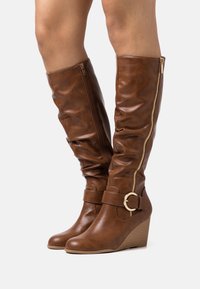 Bottes compensées hautes jusqu'au genou de couleur marron avec fermeture éclair latérale, bride et boucle dorée portées sur des jambes nues sur fond blanc.