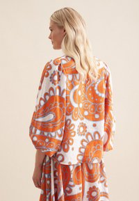 Top fluido con un vivace motivo paisley arancione e blu su tessuto bianco. Design largo con spalle abbassate e maniche ampie.