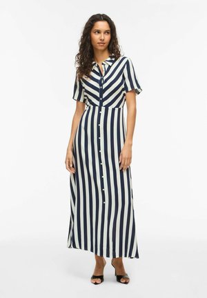 Kvinne iført en marineblå og hvit vertikalstripet skjortekjole med korte ermer og sorte åpne hæler, stående på hvit bakgrunn.