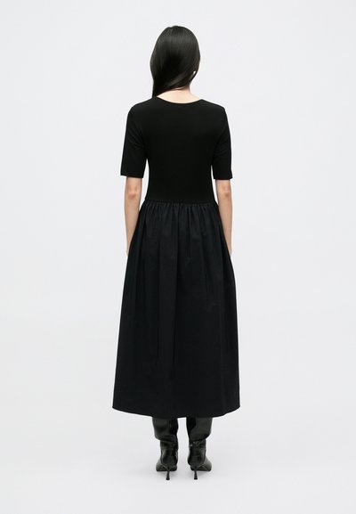 Čierne šaty s priliehavým živôtikom a voľnou, nariasenou sukňou. Krátke rukávy, okrúhly výstrih a nosené s čiernymi čižmami po kolená.