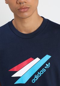 T-shirt di cotone blu navy con un grande logo multicolore di Adidas, caratterizzato da strisce angolate rosse, bianche e blu, e un collo rotondo.