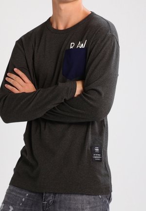 Personne portant une chemise à manches longues gris foncé avec une poche poitrine bleu marine et un petit écusson près de l'ourlet, bras croisés, debout devant un fond uni.