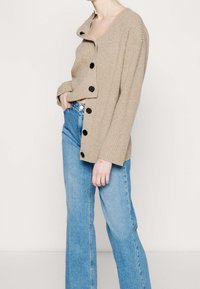 Personne portant un cardigan beige boutonné et un jean bleu clair taille haute, main dans la poche, debout devant un fond uni.