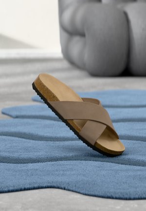 Sandalia individual de color marrón claro con tiras cruzadas y suela de corcho, descansando sobre una alfombra azul texturizada, con un gran objeto gris desenfocado en el fondo.