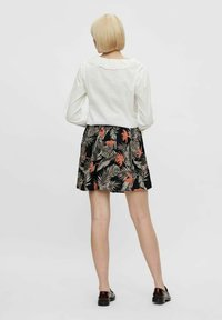 Blonďatá žena stojící zády k fotoaparátu, oblečená v bílé blůze s dlouhým rukávem a vlnitým límečkem, černé květinové sukni a tmavých mokasínech.