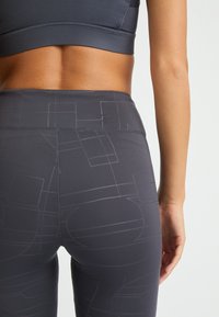 Leggings de cintura alta en gris oscuro con una textura suave, que presentan un patrón geométrico en un gris más claro y una banda elástica en la cintura.