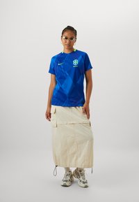 Blue football jersey with geometric patterns, Nike logo, and Brazil emblem. Paired with a beige long skirt and multi-color sneakers.