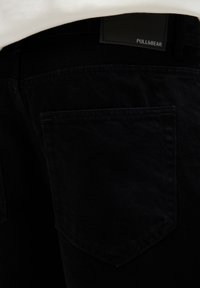 Gros plan de la poche arrière d’un jean en denim noir avec une étiquette de la marque Pull&Bear visible sur la ceinture contre un tissu blanc.