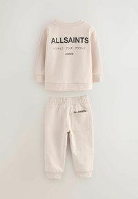 Helles beige Baumwoll-Sweatshirt und Jogginghose Set. Das Sweatshirt hat ein schwarzes Logo auf der Rückseite; die Jogginghose verfügt über eine Gesäßtasche mit passendem Logo.