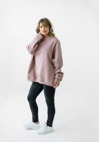 Przesadny różowy sweter z miękkiego materiału, z długimi rękawami i ściągaczami na mankietach, zestawiony z czarnymi obcisłymi dżinsami i białymi trampkami.