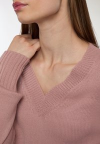 Jasnoróżowy sweter z dzianiny z dekoltem w kształcie litery V, żebrowanymi mankietami i miękką teksturą. Odcień skóry widoczny, ręka delikatnie oparta na szyi.