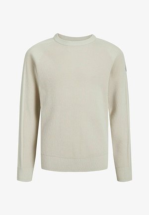 Dziecko stojące z rękami w kieszeniach, ubrane w beżowy sweter, czarne spodnie i czarne sneakersy z białymi podeszwami na jednolitym tle.