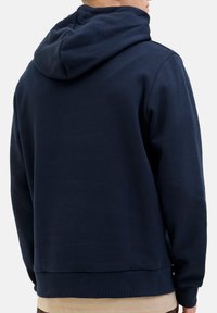 Mand iført en marineblå hættetrøje set fra ryggen, med en beige skjorte synlig nedenunder.