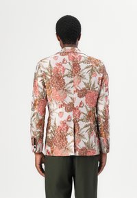 Floraler Blazer in Creme mit pinken und orangefarbenen Mustern. Mit langen Ärmeln, Reverskragen und geknöpften Manschetten. Kombiniert mit dunkelgrünen Hosen.