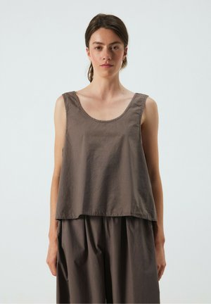 Donna con capelli scuri legati in una coda bassa indossa un top marrone senza maniche e pantaloni larghi abbinati su uno sfondo grigio semplice.