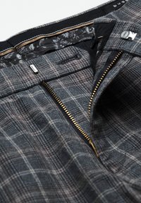 Pantalon à carreaux gris avec une fermeture éclair dorée ouverte, une fermeture métallique à crochet et barre, un bouton noir et une doublure intérieure fleurie à la ceinture.