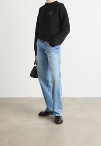 Pull noir côtelé avec logo bleu, jean bleu clair à jambes larges, et chaussures en cuir noir. Un petit sac à main noir complète le look.