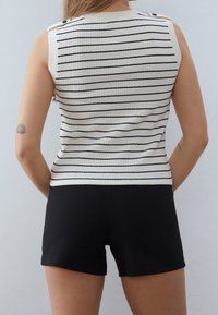 Kobieta ubrana w bezrękawnik w biało-czarne poziome paski oraz czarne szorty, stoi tyłem do kamery.
