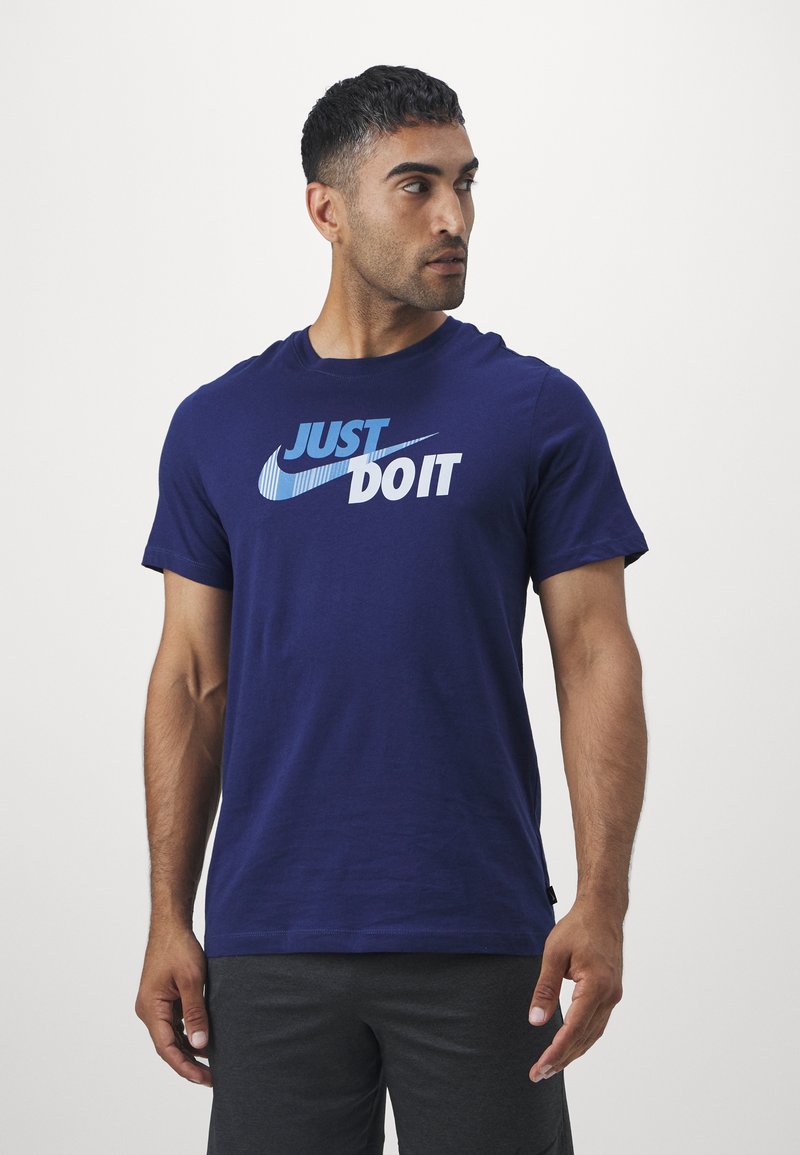 Man wearing navy blue "Just Do It" Nike t-shirt and dark shorts, looking to his left against plain white background.