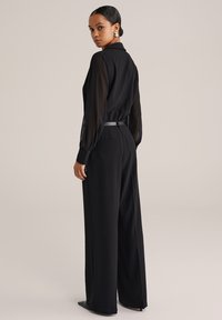 Čierny outfit pozostávajúci z priesvitnej blúzky s dlhým rukávom a golierom a vysokými nohavicami so širokými nohavicami. Obsahuje tenký opasok a zadné vrecká.