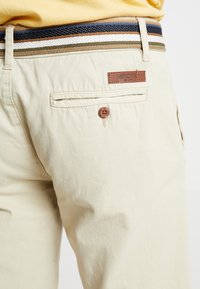 Pantaloni di cotone beige con una tasca posteriore, dotati di un'etichetta in pelle marrone del marchio e di una cintura intrecciata multicolore con strisce diagonali.