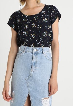 Kobieta ubrana w czarną, kwiecistą bluzkę z krótkim rękawem, włożoną do jasnoniebieskiej spódnicy dżinsowej z wysokim stanem, z przednim rozcięciem i przetarciami.