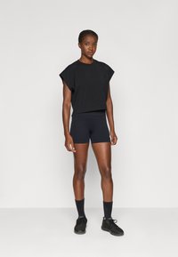 Schwarzes kurz geschnittenes T-Shirt mit weiten Ärmeln, kombiniert mit schwarzen enganliegenden Shorts. Das Model trägt schwarze Sneakers und Knöchelsocken vor einem neutralen Hintergrund.