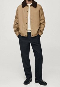 Beżowa kurtka z brązowym futrzanym kołnierzem, zapinana na zamek z dwoma bocznymi kieszeniami. Noszona na kremowej koszuli i czarnych spodniach, z butami.