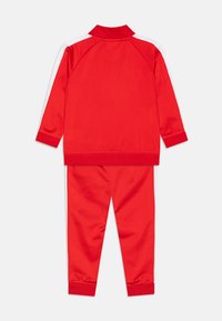 Ensemble de survêtement rouge comprenant une veste à col et un pantalon assorti. Fabriqué en tissu lisse avec des rayures blanches sur les côtés de la veste.
