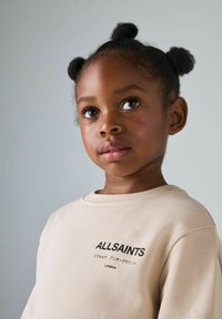 Beige Sweatshirt mit Rundhalsausschnitt, versehen mit dem "ALLSAINTS"-Logo in schwarzem Text. Das Modell hat kurzes, strukturiertes Haar, das zu zwei Duttfrisuren gestylt ist.
