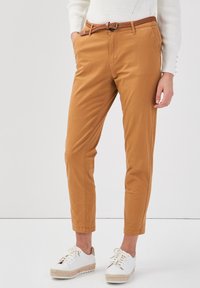 Pantalons en coton beige avec une ceinture tressée marron, de coupe slim et avec des poignets. Portés avec des baskets blanches à semelle texturée.
