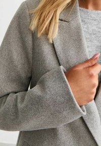 Blazer gris clair en mélange de laine avec un col cranté, une texture douce, et des manches légèrement surdimensionnées ; une main est visible près de la zone du col.