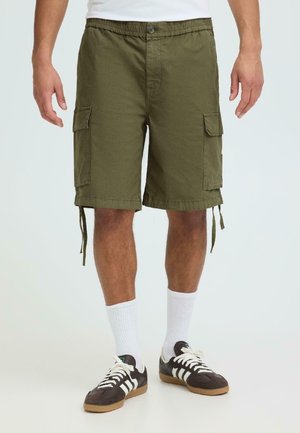 Mann trägt olivgrüne Cargo-Shorts mit Seitentaschen, weiße Sportsocken und schwarz-weiße Turnschuhe und steht vor einem schlichten Hintergrund.
