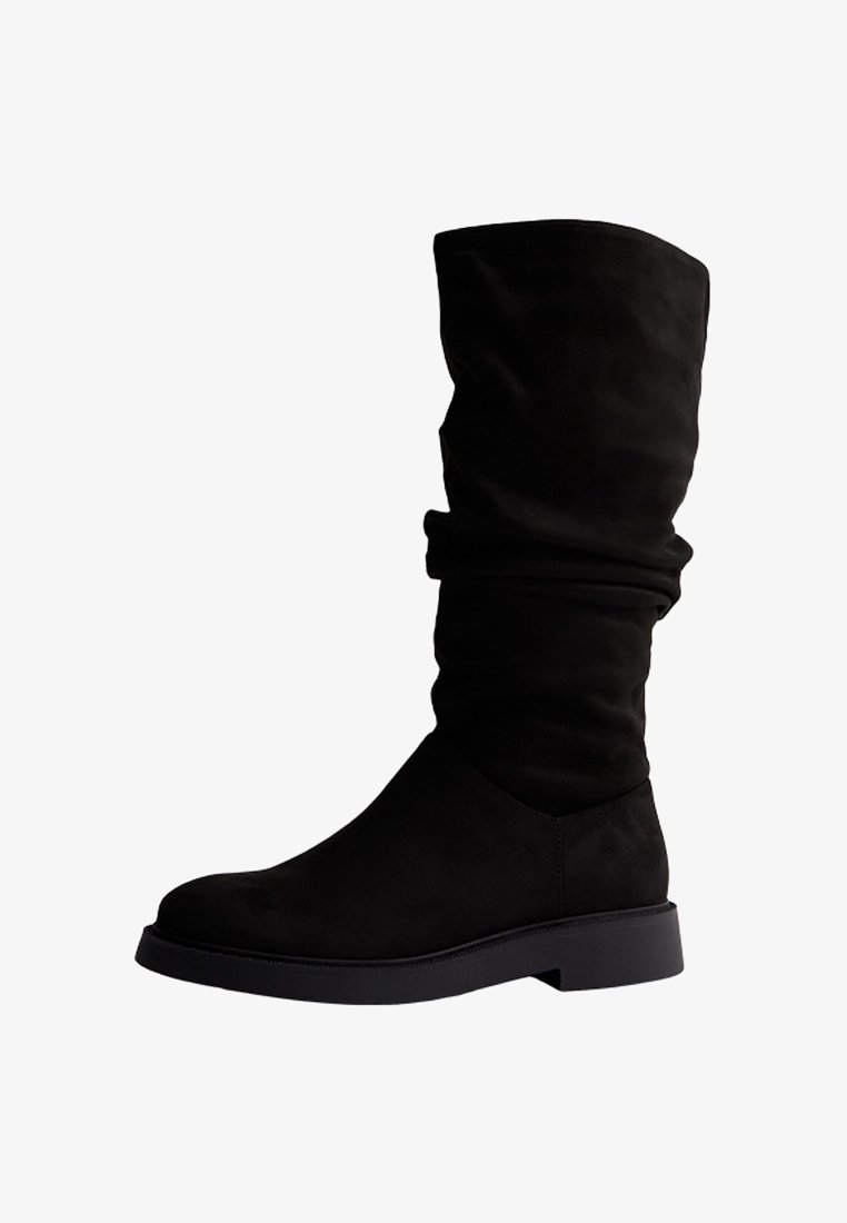 Botte noire en daim mi-mollet avec semelle plate et talon léger, présentant un design supérieur relâché et un bout arrondi.