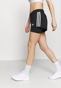 Osoba kráčející v černých šortkách Adidas s bílými pruhy, bílých ponožkách a bílých sportovních botách na jednobarevném bílém pozadí.