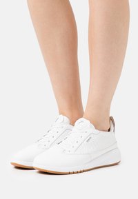 Zapatillas deportivas blancas con paneles perforados, cordones redondos y un collar acolchado. Suela de goma en un color marrón contraste.