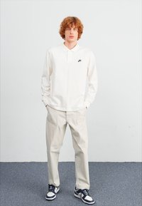 Polo branco de manga longa com gola e logótipo preto da Nike, combinado com calças beje claras e ténis azuis e brancos.