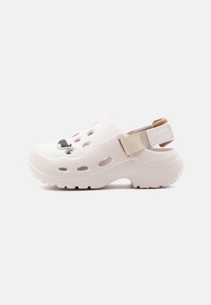 Sabot blanc à enfiler avec des trous de ventilation, une bride de talon beige réglable et une petite décoration en forme de baleine sur le dessus avant.
