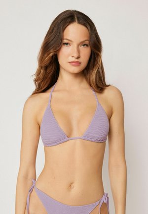 Ung kvinde med langt brunt hår iført en tekstureret lavendel trekants-bikinitop og matchende side-bindebikinibund mod en ensfarvet baggrund.