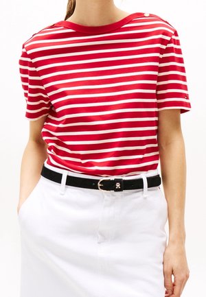 Osoba ubrana w czerwono-białą koszulkę w paski z krótkim rękawem, włożoną do białej spódnicy z czarnym paskiem, z rękami w kieszeniach spódnicy.