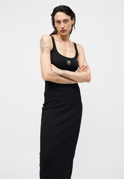 Γυναίκα με τατουάζ και σκουλαρίκια, ντυμένη με μαύρο αμάνικο φόρεμα, όρθια με σταυρωμένα τα χέρια μπροστά σε απλό φόντο.