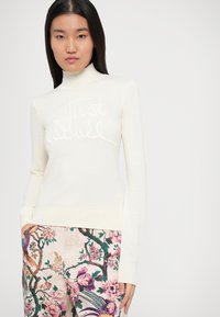 Kremasta pulover s turtleneck ovratnikom, ki ima strukturiran dizajn "Just Natural" na sprednji strani, v kombinaciji z vzorčenimi cvetličnimi hlačami v živahnih barvah.