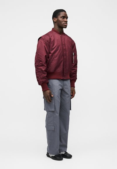 Bordová bomber bunda s rebrovanými manžetami a vreckom na zips, doplnená sivými cargo nohavicami so bočnými vreckami a voľným strihom.