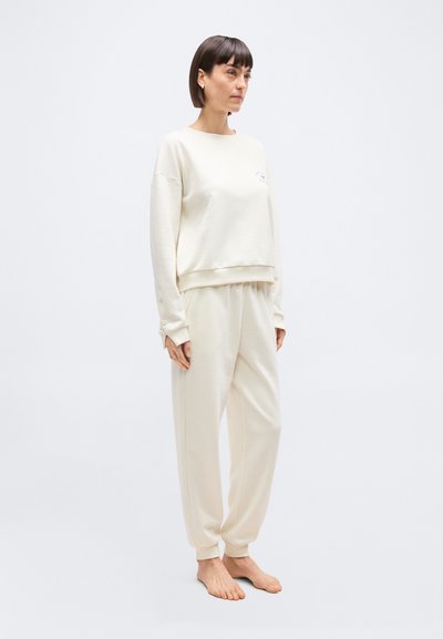 Γυναίκα που στέκεται ξυπόλυτη, φορώντας εκρού φούτερ με μακριά μανίκια και ταιριαστή φόρμα, μπροστά σε απλό ανοιχ τό φόντο.