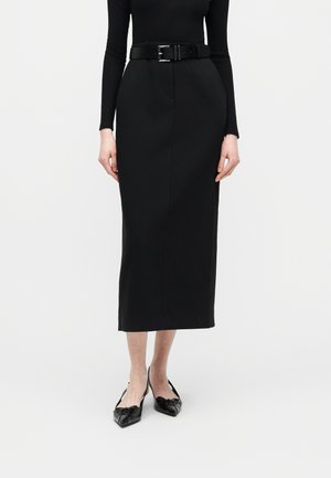 Γυναίκα που φοράει μαύρο εφαρμοστό τοπ με μακριά μανίκια, μπηγμένο σε μαύρη midi φούστα με ψηλή μέση, μαύρη ζώνη και μαύρες μυτερές μπαλαρίνες.