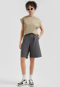 Chemise beige à manches courtes avec un petit logo, shorts gris cintrés, chaussettes blanches et baskets noir et blanc. Modèle portant des lunettes de soleil.