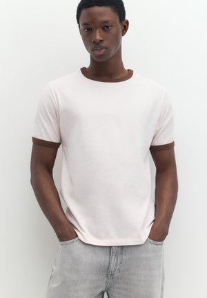 Jeune homme debout, les mains dans les poches, portant un t-shirt blanc avec un col et des bords de manches marron et un jean gris clair.