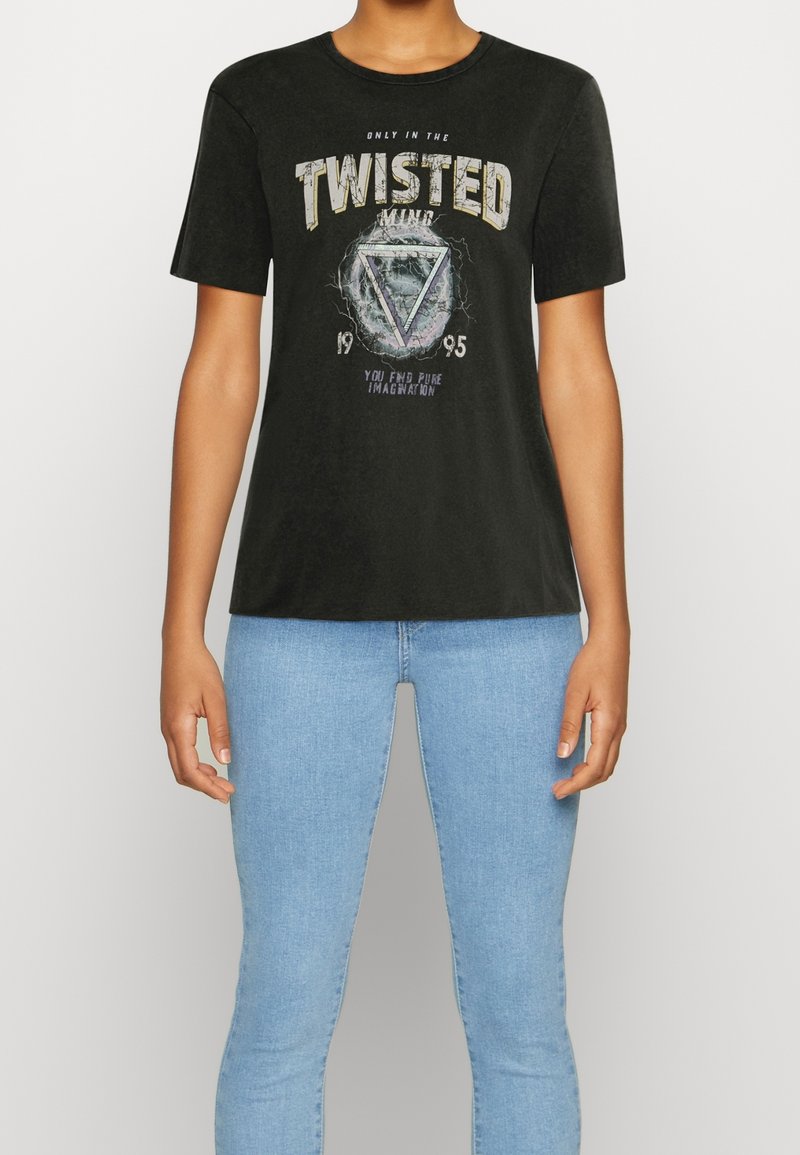 Personne portant un T-shirt noir avec le texte « TWISTED MIND » et un graphique en forme de triangle, associé à un jean bleu clair, sur un fond uni.